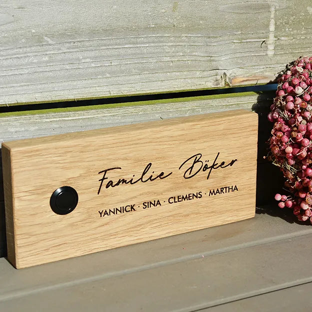 Klingelschild aus Eiche in der Größe 20 cm x 9 cm Querformat mit schwarzem Aluminiumtaster linksseitig des Familiennamens. Der Nachname ist in einer geschwungenen Handschrift graviert dazu die Vornamen in 2. Zeile in Großbuchstaben und Druckschrift.