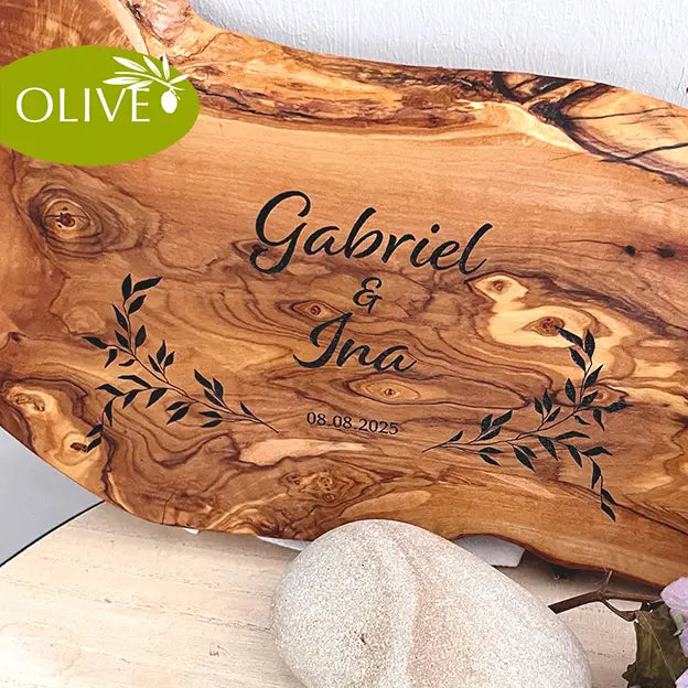 Glattes Schneidebrett aus Olivenholz mit Naturrand und personalisierter Gravur Motiv Reigen mit Namen und Datum in in modernen geschwungenen Schrift. Hier als Frühstücksbrett oder glattes Schneidebrett mit Paarnamen untereinander