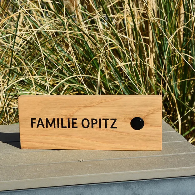 Klingelschild aus Eiche in der Größe 24 cm x 9 cm Querformat mit schwarzem Aluminiumtaster rechtsseitig des Familiennamens. Der Nachname ist in Großbuchstaben und Druckschrift.