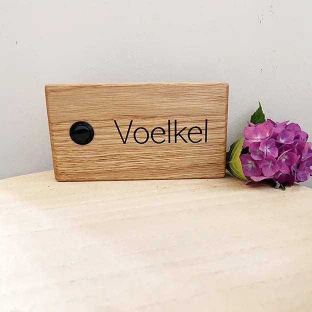 Klingelschild aus Eiche in der Größe 16 cm x 9 cm Querformat mit schwarzem Aluminiumtaster linksseitig des Familiennamens. Der Nachname ist in Groß- und Kleinschreibung in Druckschrift.
