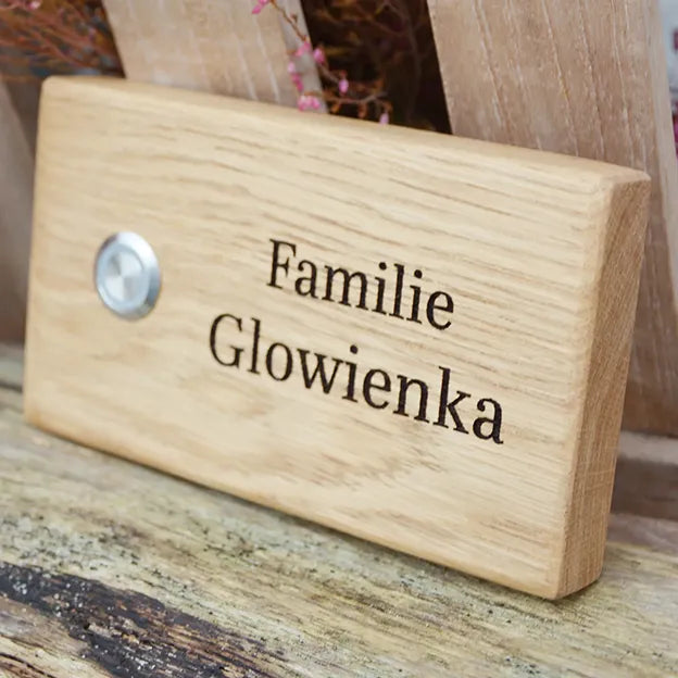 Klingelschild aus Eiche in der Größe 16 cm x 9 cm Querformat mit LED-Edelstahltaster matt linksseitig des Familiennamens. Der Nachname mit dem Wort Familie davor ist in einer klassischen Serifenschrift in 2 Zeilen graviert.