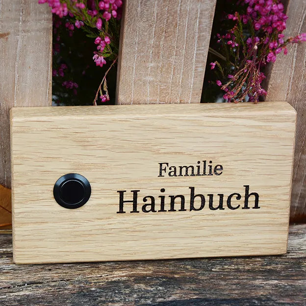 Klingelschild aus Eiche in der Größe 16 cm x 9 cm Querformat mit schwarzem Aluminiumtaster linksseitig des Familiennamens. Der Nachname ist in einer geschwungenen Handschrift graviert.