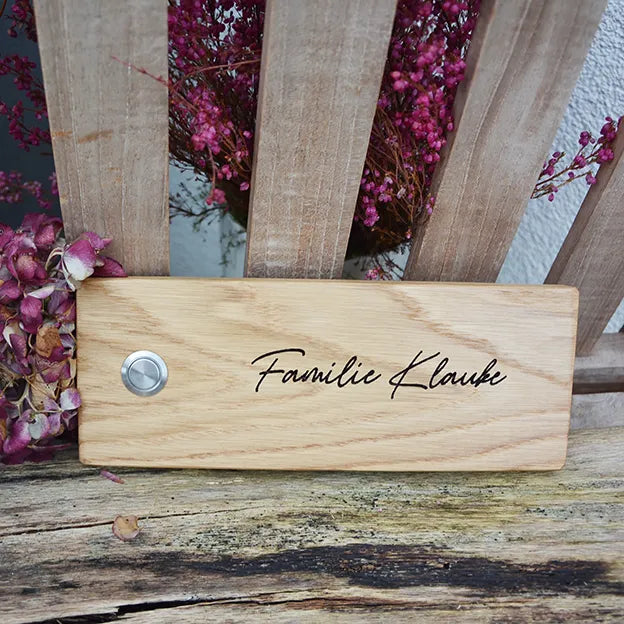 Klingelschild aus Eiche in der Größe 24 cm x 9 cm Querformat mit Edelstahltaster matt linksseitig des Familiennamens. Der Nachname ist in einer geschwungenen Handschrift graviert.