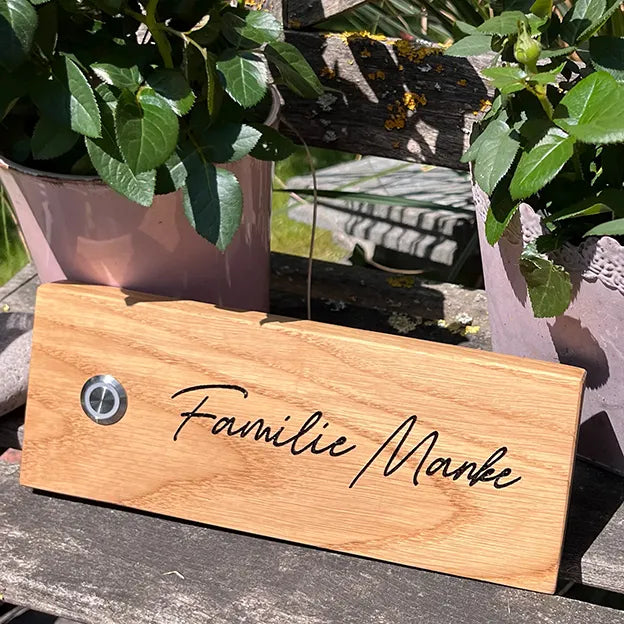 Klingelschild aus Eiche in der Größe 20 cm x 9 cm Querformat mit LED-Edelstahltaster matt linksseitig des Familiennamens. Der Nachname ist in einer geschwungenen Handschrift graviert.