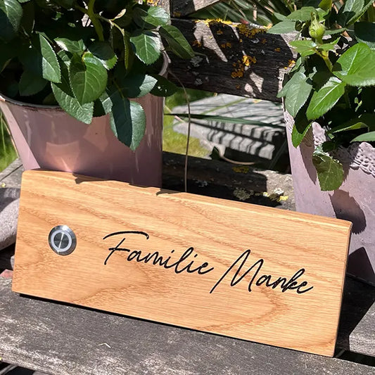 Klingelschild aus Eiche in der Größe 20 cm x 9 cm Querformat mit LED-Edelstahltaster matt linksseitig des Familiennamens. Der Nachname ist in einer geschwungenen Handschrift graviert.