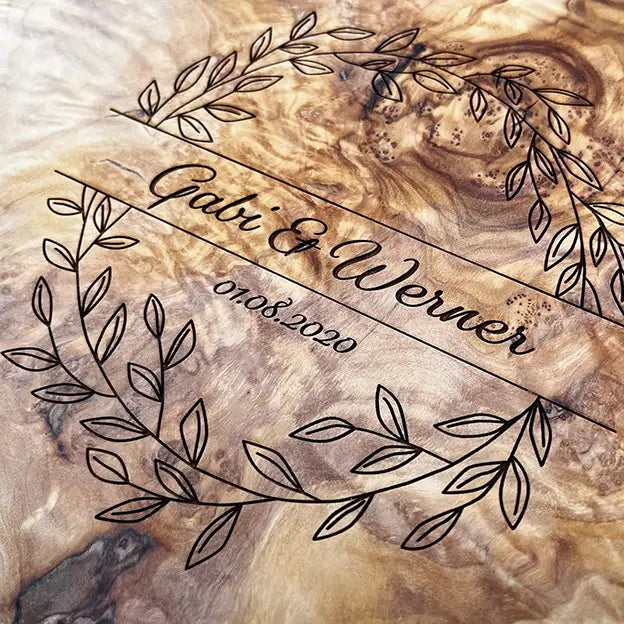 Glattes Schneidebrett aus Olivenholz mit Naturrand und personalisierter Gravur Motiv Blätterkranz mit Namen und Datum in in geschwungener Schrift. Hier als Frühstücksbrett oder glattes Schneidebrett in verschiedenen Größen und Brettformen als Hochzeitsgeschenk oder zum Jahrestag