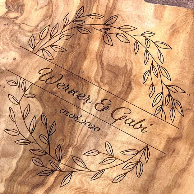 Glattes Schneidebrett aus Olivenholz mit Naturrand und personalisierter Gravur Motiv Blätterkranz mit Namen und Datum in in geschwungener Schrift. Hier als Frühstücksbrett oder glattes Schneidebrett in verschiedenen Größen und Brettformen