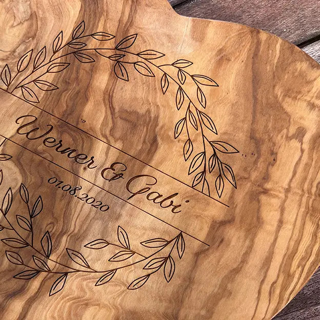 Glattes Schneidebrett aus Olivenholz mit Naturrand und personalisierter Gravur Motiv Blätterkranz mit Namen und Datum in in geschwungener Schrift. Hier als Frühstücksbrett in Herzform oder glattes Schneidebrett in verschiedenen Größen und Brettformen zum Jubiläum oder Hochzeit