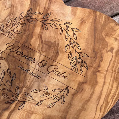 Glattes Schneidebrett aus Olivenholz mit Naturrand und personalisierter Gravur Motiv Blätterkranz mit Namen und Datum in in geschwungener Schrift. Hier als Frühstücksbrett in Herzform oder glattes Schneidebrett in verschiedenen Größen und Brettformen zum Jubiläum oder Hochzeit