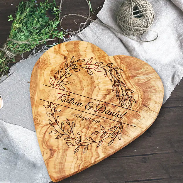 Glattes Schneidebrett aus Olivenholz mit Naturrand und personalisierter Gravur Motiv Blätterkranz mit Namen und Datum in in geschwungener Schrift. Hier als Frühstücksbrett in Herzform oder glattes Schneidebrett in verschiedenen Größen und Brettformen