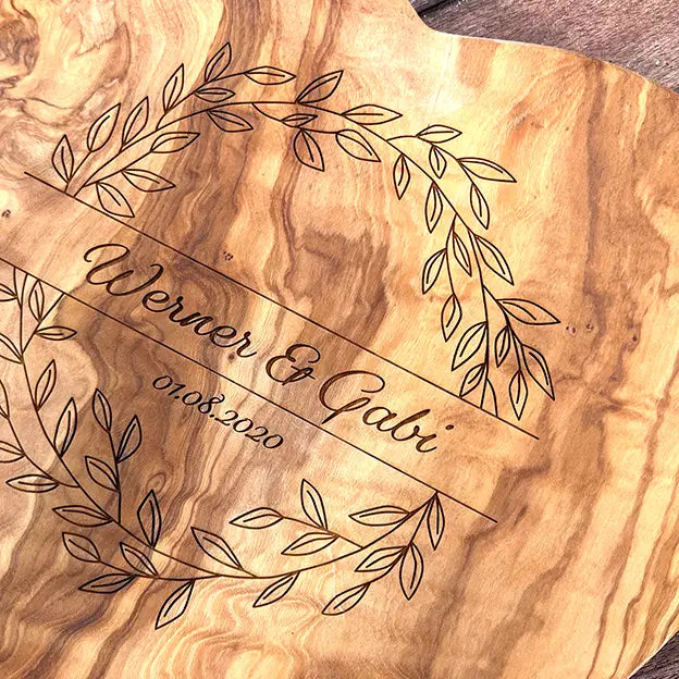 Glattes Schneidebrett aus Olivenholz mit Naturrand und personalisierter Gravur Motiv Blätterkranz mit Namen und Datum in in geschwungener Schrift. Hier als Frühstücksbrett in Herzform oder glattes Schneidebrett in verschiedenen Größen und Brettformen