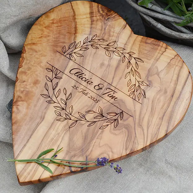 Glattes Schneidebrett aus Olivenholz mit Naturrand und personalisierter Gravur Motiv Blätterkranz mit Namen und Datum in in geschwungener Schrift. Hier als Frühstücksbrett in Herzform oder glattes Schneidebrett in verschiedenen Größen und Brettformen zur Verlobung oder Jahrestag