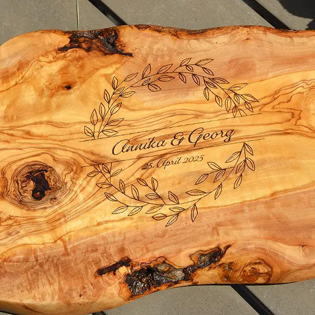 Glattes Schneidebrett aus Olivenholz mit Naturrand und personalisierter Gravur Motiv Blätterkranz mit Namen und Datum in in geschwungener Schrift. Hier als Frühstücksbrett oder glattes Schneidebrett in verschiedenen Größen und Brettformen