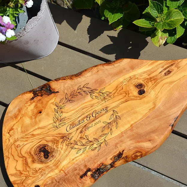 Glattes Schneidebrett aus Olivenholz mit Naturrand und personalisierter Gravur Motiv Blätterkranz mit Namen und Datum in in geschwungener Schrift. Hier als Frühstücksbrett mit Handgriff oder glattes Schneidebrett in verschiedenen Größen und Brettformen