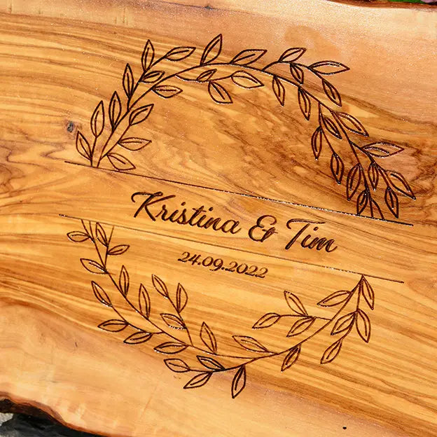 Glattes Schneidebrett aus Olivenholz mit Naturrand und personalisierter Gravur Motiv Blätterkranz mit Namen und Datum in in geschwungener Schrift. Hier als Frühstücksbrett oder glattes Schneidebrett in verschiedenen Größen und Brettformen zur Silberhochzeit oder zum Jahrestag