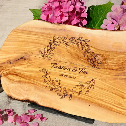 Glattes Schneidebrett aus Olivenholz mit Naturrand und personalisierter Gravur Motiv Blätterkranz mit Namen und Datum in in geschwungener Schrift. Hier als Frühstücksbrett oder glattes Schneidebrett in verschiedenen Größen und Brettformen
