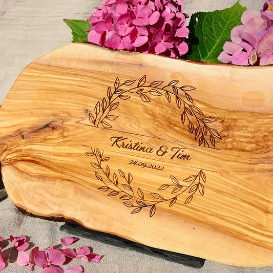 Glattes Schneidebrett aus Olivenholz mit Naturrand und personalisierter Gravur Motiv Blätterkranz mit Namen und Datum in in geschwungener Schrift. Hier als Frühstücksbrett oder glattes Schneidebrett in verschiedenen Größen und Brettformen