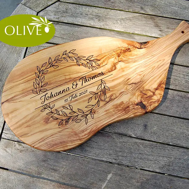 Glattes Schneidebrett aus Olivenholz mit Naturrand und personalisierter Gravur Motiv Blätterkranz mit Namen und Datum in in geschwungener Schrift. Hier als Frühstücksbrett mit Handgriff oder glattes Schneidebrett in verschiedenen Größen und Brettformen
