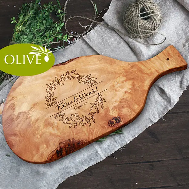 Glattes Schneidebrett aus Olivenholz mit Naturrand und personalisierter Gravur Motiv Blätterkranz mit Namen und Datum in in geschwungener Schrift. Hier als Frühstücksbrett mit Handgriff oder glattes Schneidebrett in verschiedenen Größen und Brettformen