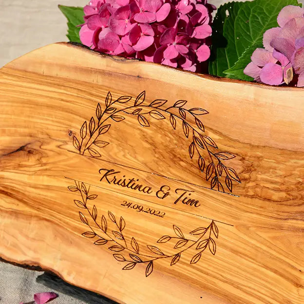 Glattes Schneidebrett aus Olivenholz mit Naturrand und personalisierter Gravur Motiv Blätterkranz mit Namen und Datum in in geschwungener Schrift. Hier als Frühstücksbrett oder glattes Schneidebrett in verschiedenen Größen und Brettformen