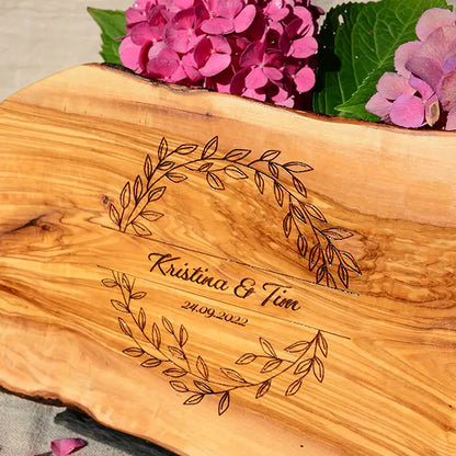 Glattes Schneidebrett aus Olivenholz mit Naturrand und personalisierter Gravur Motiv Blätterkranz mit Namen und Datum in in geschwungener Schrift. Hier als Frühstücksbrett oder glattes Schneidebrett in verschiedenen Größen und Brettformen