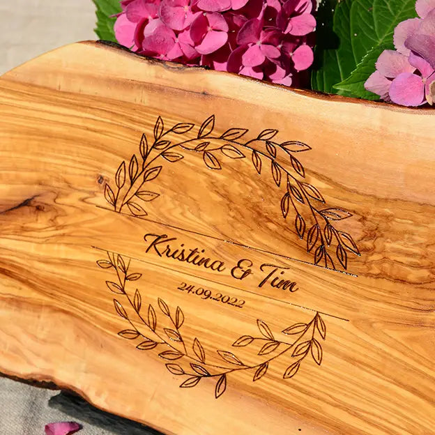 Glattes Schneidebrett aus Olivenholz mit Naturrand und personalisierter Gravur Motiv Blätterkranz mit Namen und Datum in in geschwungener Schrift. Hier als Frühstücksbrett oder glattes Schneidebrett in verschiedenen Größen und Brettformen
