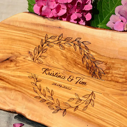 Glattes Schneidebrett aus Olivenholz mit Naturrand und personalisierter Gravur Motiv Blätterkranz mit Namen und Datum in in geschwungener Schrift. Hier als Frühstücksbrett oder glattes Schneidebrett in verschiedenen Größen und Brettformen