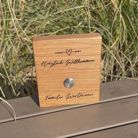 Klingelschild aus edler Eiche mit Edelstahl Taster unbeleuchtet oder mit weißem LED-Licht oder schwarzen Aluminium-Taster in der Größe 12 x 14 cm oder 14 x 25 cm. Verspieltes Motiv mit einem Herz und vier Kreisen auf einer Linie mit Herzlich Willkommen und Deinem Nachnamen - optional mit Vornamen oder “Familie” - wird per Laser in das Holz graviert. Schriftart: Geschwungene Handschrift. Hier mit Edelstahltaster matt.